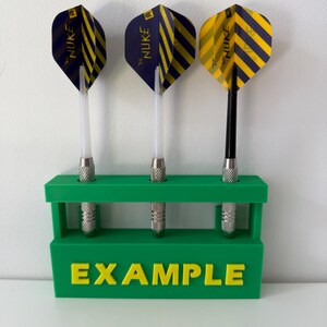 May include: Three darts with striped blue and yellow flights are displayed in a green holder. The darts have silver tips and clear shafts. The holder has the word "EXAMPLE" in yellow letters.