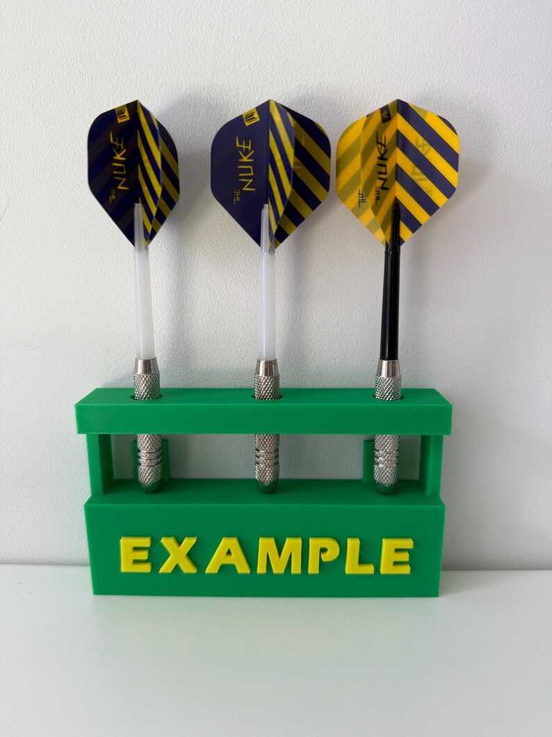 May include: Three darts with striped blue and yellow flights are displayed in a green holder. The darts have silver tips and clear shafts. The holder has the word "EXAMPLE" in yellow letters.