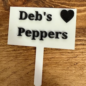 Puede incluir: Un marcador de jard&iacute;n blanco con el texto "Deb's Peppers" en negro y un s&iacute;mbolo de coraz&oacute;n negro. El marcador est&aacute; sobre una superficie de madera.