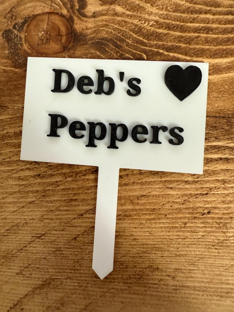 Puede incluir: Un marcador de jard&iacute;n blanco con el texto "Deb's Peppers" en negro y un s&iacute;mbolo de coraz&oacute;n negro. El marcador est&aacute; sobre una superficie de madera.