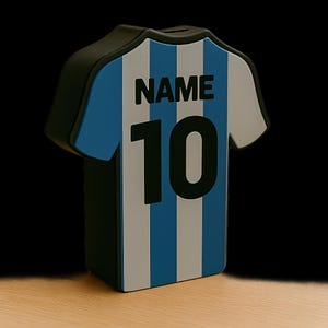 K&ouml;nnte beinhalten: Ein blau-wei&szlig;-schwarzes Sparschwein in Form eines Fu&szlig;balltrikots. Das Trikot hat vertikale blaue und wei&szlig;e Streifen mit der Nummer "10" und dem Wort "NAME" auf der R&uuml;ckseite. Das Sparschwein besteht aus einem harten Material und steht auf einer Holzoberfl&auml;che.