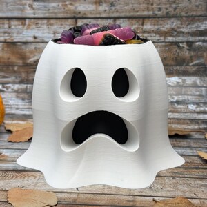 May include: A white ghost-shaped candy bowl with a wide, open mouth and two oval eyes. The bowl is filled with colourful gummy sweets. A small orange pumpkin and autumn leaves are in the background.