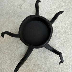 May include: Black decorative bowl with six curved legs. The bowl has a textured interior and a detailed rim. The legs are curved and end in a foot-like shape. The object is made of a dark material.