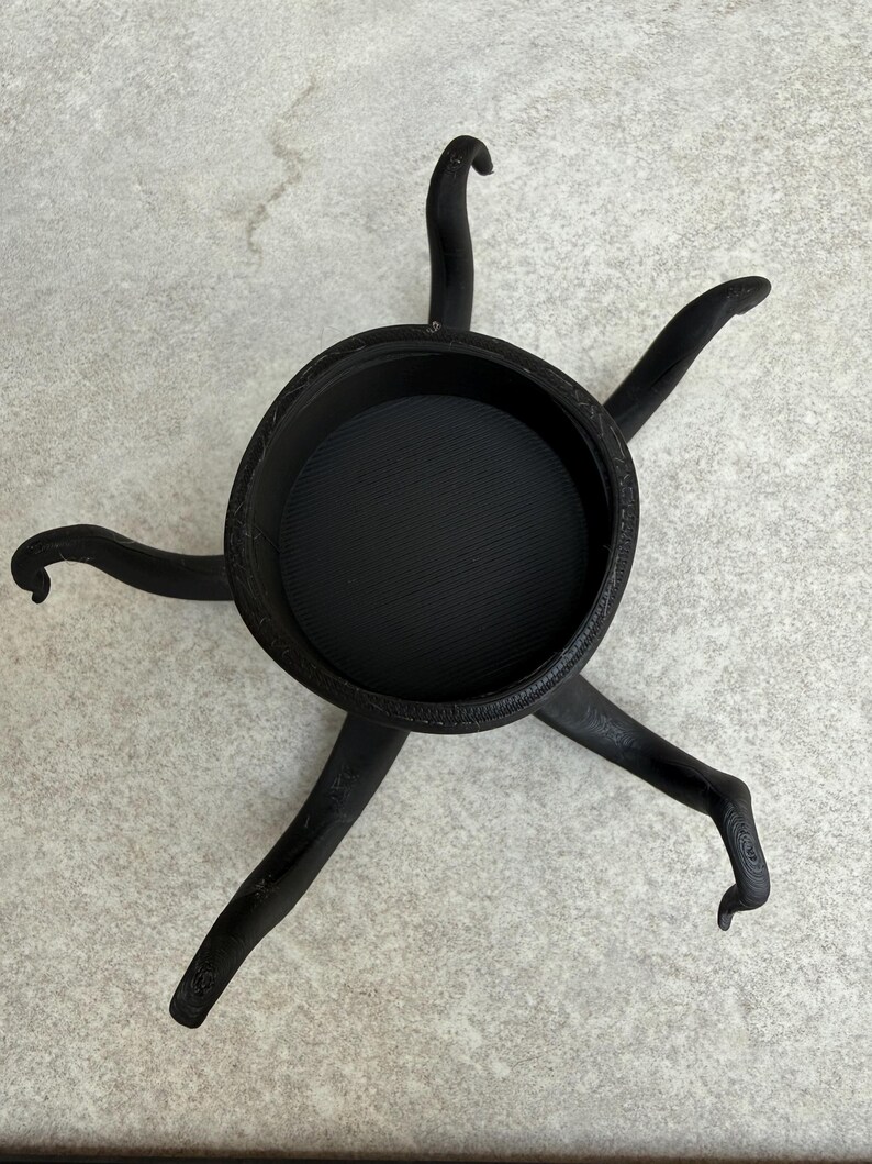 May include: Black decorative bowl with six curved legs. The bowl has a textured interior and a detailed rim. The legs are curved and end in a foot-like shape. The object is made of a dark material.