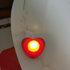 May include: A red heart-shaped LED light with a bright yellow center. The light is placed on a white surface with a floral design. The light is a decorative item.