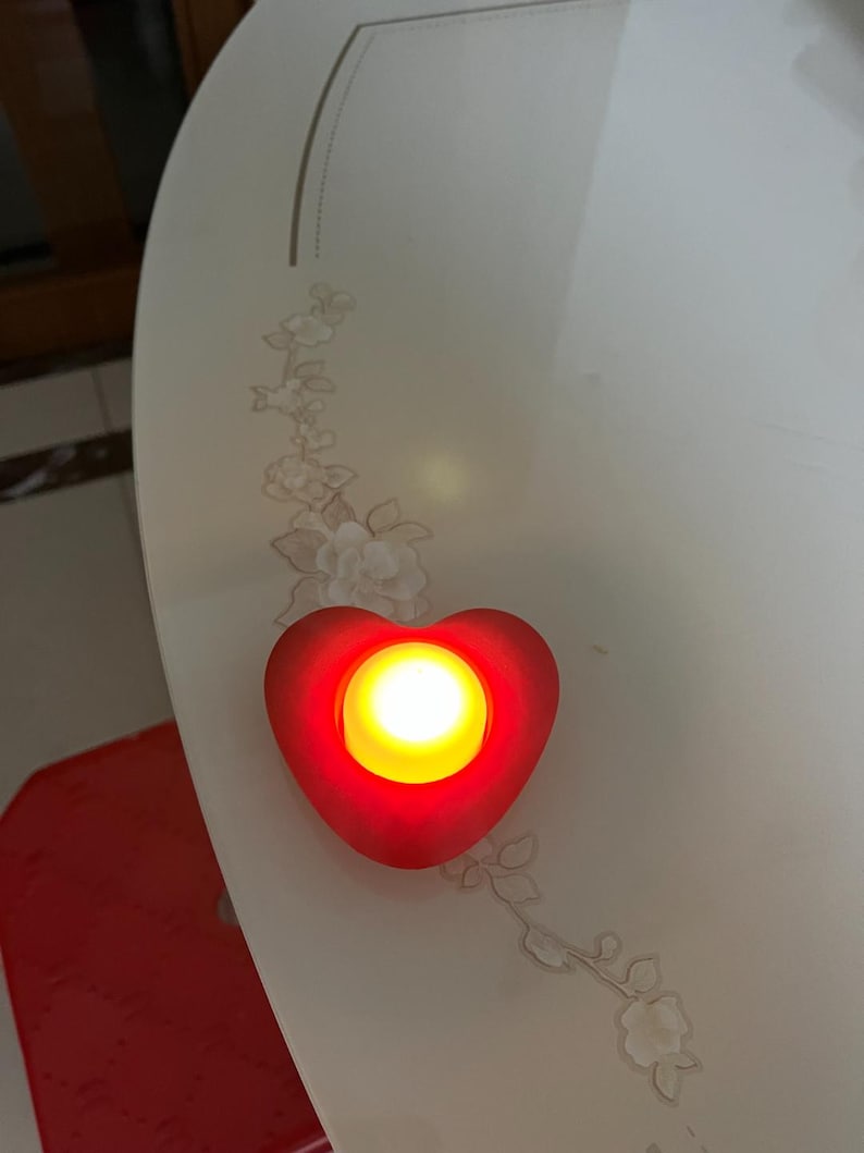 May include: A red heart-shaped LED light with a bright yellow center. The light is placed on a white surface with a floral design. The light is a decorative item.