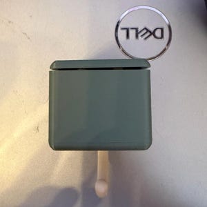 May include: A sage green, rectangular object with a small, rounded peg extending from the bottom. The object has a lid and is positioned near a silver Dell logo.