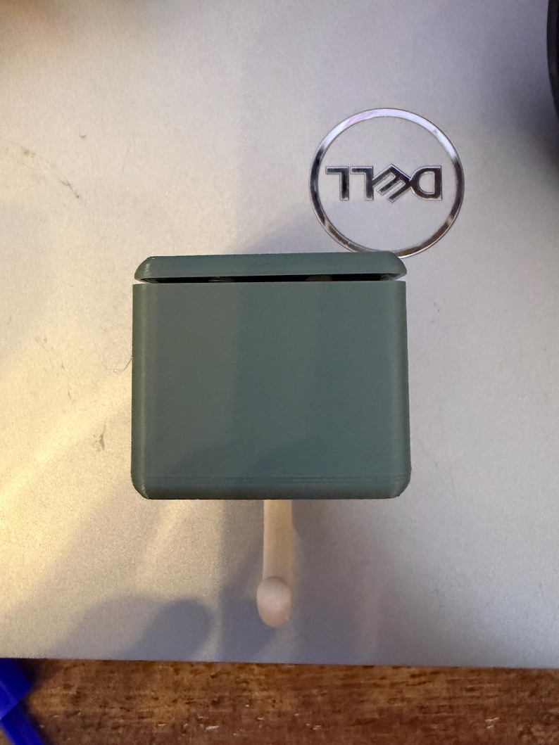 May include: A sage green, rectangular object with a small, rounded peg extending from the bottom. The object has a lid and is positioned near a silver Dell logo.