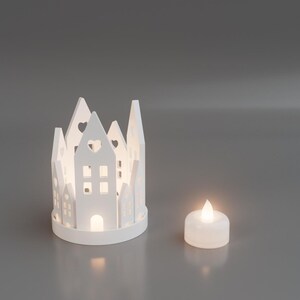 May include: A white, decorative candle holder shaped like a village, with cut-out windows and heart-shaped details. A small, lit, white candle sits next to the village. The scene is set against a gray background.