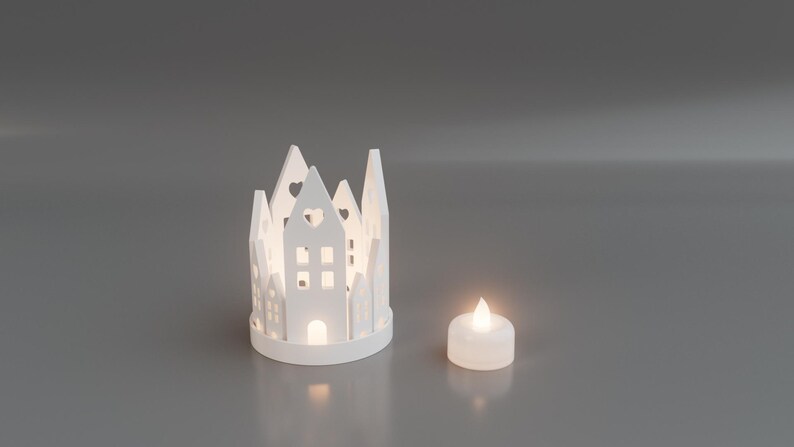 May include: A white, decorative candle holder shaped like a village, with cut-out windows and heart-shaped details. A small, lit, white candle sits next to the village. The scene is set against a gray background.