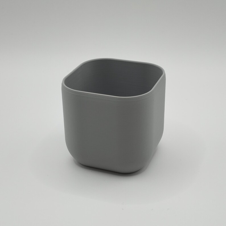 May include: A gray, square-shaped container with rounded corners. The container is made of a solid material and has a smooth, matte finish. The interior is a slightly darker shade of gray. The container is empty.