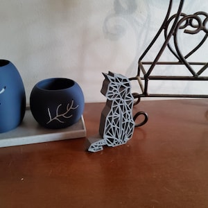 May include: Two navy blue decorative vases with white branch designs sit on a gray tray. A geometric gray cat figurine is next to them. A wrought iron decorative stand is in the background. The objects are on a brown surface.