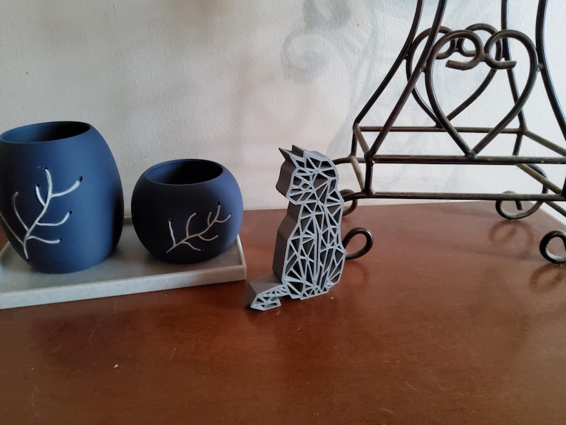 May include: Two navy blue decorative vases with white branch designs sit on a gray tray. A geometric gray cat figurine is next to them. A wrought iron decorative stand is in the background. The objects are on a brown surface.