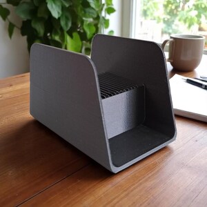 May include: A dark gray desk organizer with a rectangular base and curved sides. The organizer has a section with horizontal slats. The item is sitting on a wooden surface.