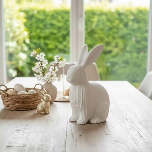 May include: A white ceramic rabbit figurine with long ears sits on a wooden table. A small gold glitter rabbit, a basket of eggs, and a vase of white flowers are also on the table. The background includes a window and greenery.