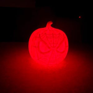 May include: A glowing red pumpkin-shaped decoration with a Spider-Man mask design. The pumpkin is illuminated against a dark background, creating a spooky and festive Halloween decoration.