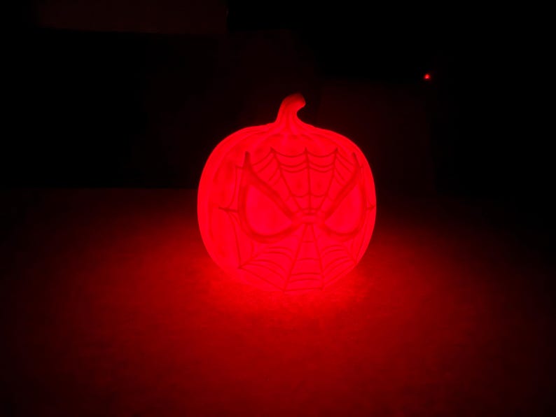 May include: A glowing red pumpkin-shaped decoration with a Spider-Man mask design. The pumpkin is illuminated against a dark background, creating a spooky and festive Halloween decoration.