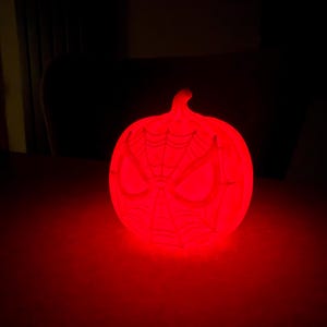 May include: A glowing red pumpkin-shaped light with a Spider-Man face design. The light is emitting a bright red glow, and the design is visible on the front of the pumpkin. The light is sitting on a dark surface.