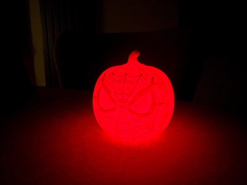 May include: A glowing red pumpkin-shaped light with a Spider-Man face design. The light is emitting a bright red glow, and the design is visible on the front of the pumpkin. The light is sitting on a dark surface.