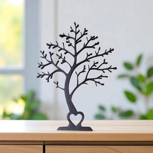 May include: A black metal tree-shaped jewelry holder with a heart cutout at the base. The tree has many branches for hanging necklaces and other jewelry. The holder is displayed on a wooden surface.