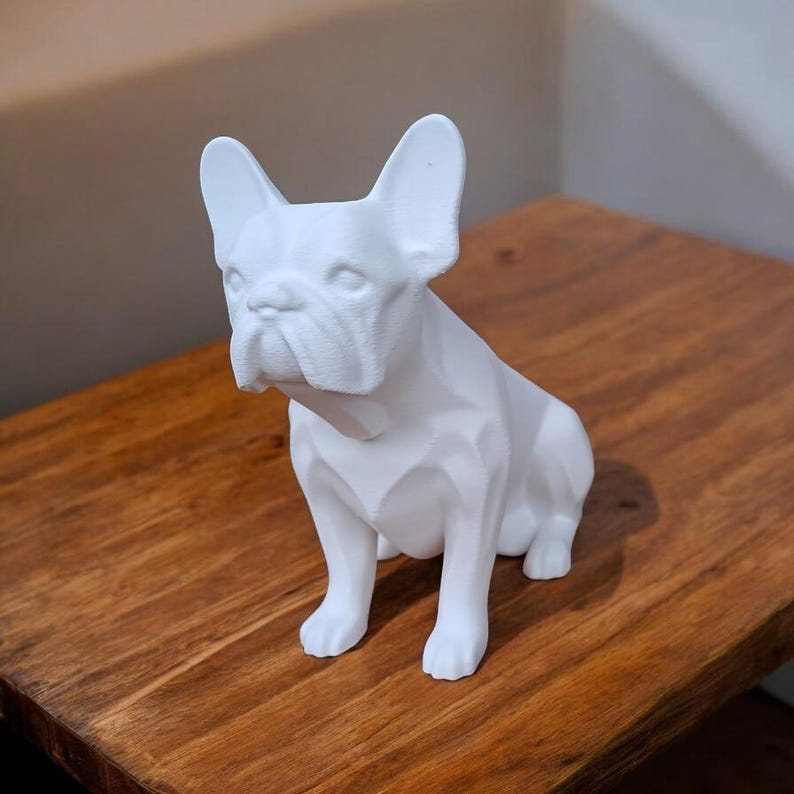 May include: White ceramic figurine of a French bulldog sitting with its legs crossed. The figurine is made in a geometric style, with a simplified, blocky design.