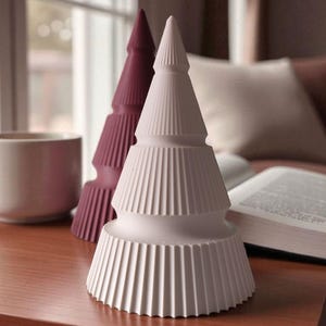 May include: Two decorative ceramic Christmas trees. One is white, the other is a deep burgundy. Both have a ridged, layered design. The trees are on a wooden surface, with a mug and open book in the background.