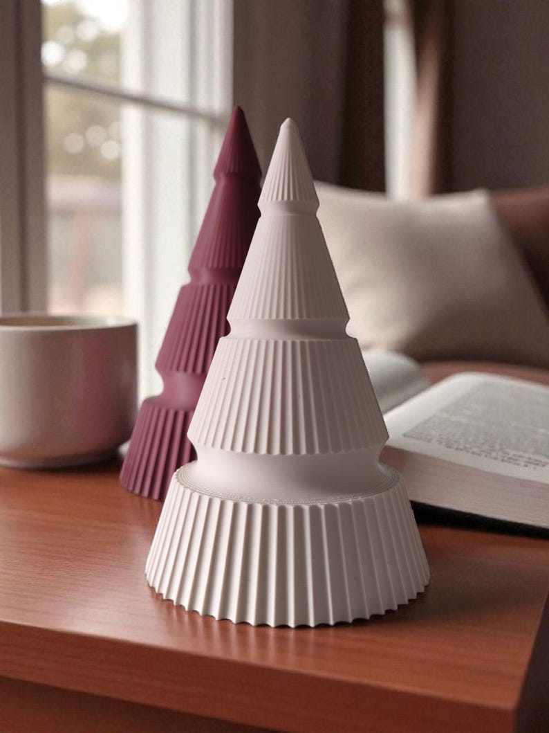 May include: Two decorative ceramic Christmas trees. One is white, the other is a deep burgundy. Both have a ridged, layered design. The trees are on a wooden surface, with a mug and open book in the background.