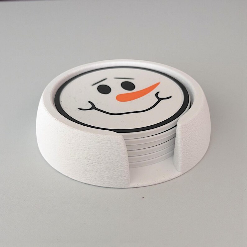 May include: A set of snowman-themed coasters with a white holder. Each coaster features a smiling snowman face with black eyes, a carrot nose, and a black outline. The holder is white and textured.