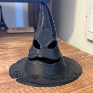 Op de afbeelding: Een zwarte, 3D-geprinte Sorteerhoed met een brede rand en een puntige top. De hoed heeft oog- en mondopeningen, waardoor hij een gezichtsuitdrukking krijgt. De hoed staat op een houten oppervlak.