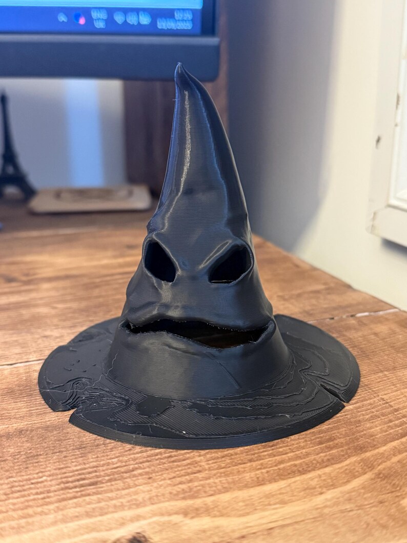 Op de afbeelding: Een zwarte, 3D-geprinte Sorteerhoed met een brede rand en een puntige top. De hoed heeft oog- en mondopeningen, waardoor hij een gezichtsuitdrukking krijgt. De hoed staat op een houten oppervlak.