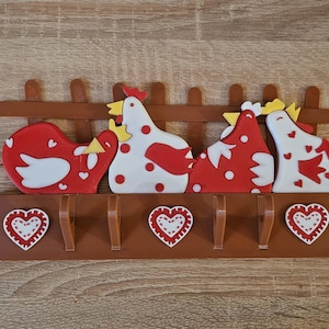 Puede incluir: Un perchero decorativo de pared con cuatro gallinas estilizadas en rojo y blanco, posadas sobre cuatro ganchos. El perchero incluye un dise&ntilde;o de valla marr&oacute;n y detalles en forma de coraz&oacute;n. Las dimensiones son de unos 30 cm de ancho y 15 cm de alto.