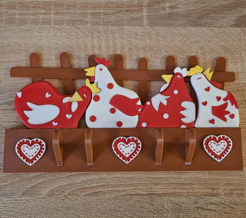Puede incluir: Un perchero decorativo de pared con cuatro gallinas estilizadas en rojo y blanco, posadas sobre cuatro ganchos. El perchero incluye un dise&ntilde;o de valla marr&oacute;n y detalles en forma de coraz&oacute;n. Las dimensiones son de unos 30 cm de ancho y 15 cm de alto.