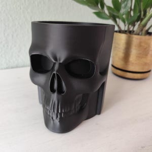 May include: A black skull-shaped planter made of a solid material. The planter has a detailed skull design with eye sockets, a nose cavity, and teeth. The planter is designed to hold small plants or succulents.