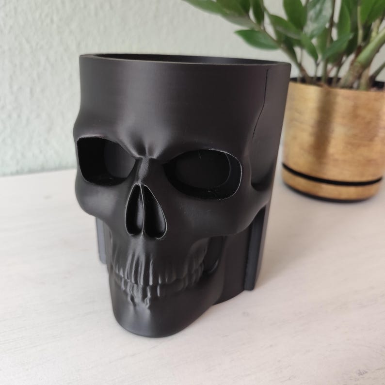 May include: A black skull-shaped planter made of a solid material. The planter has a detailed skull design with eye sockets, a nose cavity, and teeth. The planter is designed to hold small plants or succulents.