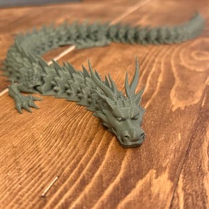 May include: A detailed, articulated dragon figurine in a muted green colour. The dragon has a spiky spine, detailed scales, and a fierce expression. The figurine is lying on a wooden surface.