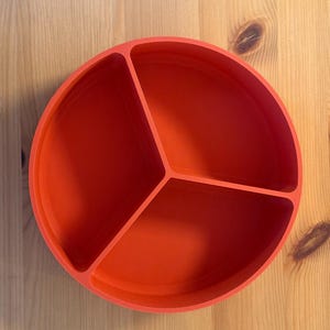 May include: A bright orange, round divided plate with three sections. The plate is made of a solid material and sits on a light-colored wooden surface. The sections are separated by raised dividers.