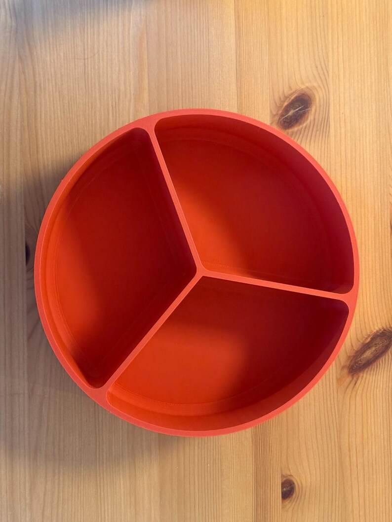 May include: A bright orange, round divided plate with three sections. The plate is made of a solid material and sits on a light-colored wooden surface. The sections are separated by raised dividers.