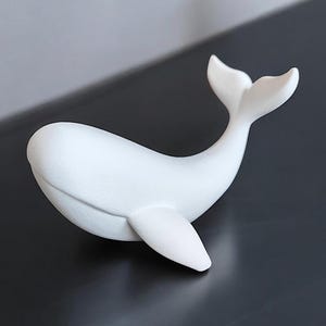 May include: A white whale figurine with a smooth, matte finish. The whale is depicted in a curved pose, with its tail raised and a single fin extended. The figurine is a decorative item.