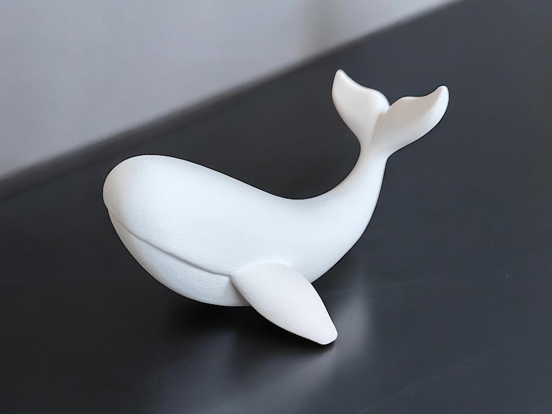 May include: A white whale figurine with a smooth, matte finish. The whale is depicted in a curved pose, with its tail raised and a single fin extended. The figurine is a decorative item.