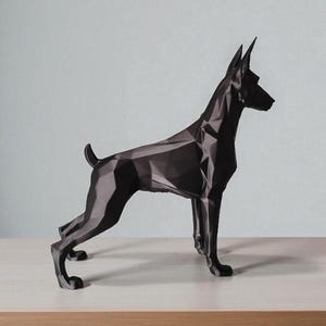 Op de afbeelding: Een zwarte geometrische sculptuur van een hond, waarschijnlijk een Dobermann, op een licht houten oppervlak. De vorm van de hond is gemaakt van hoekige vlakken, wat een moderne, abstracte uitstraling geeft. De achtergrond is effen lichtgrijs.