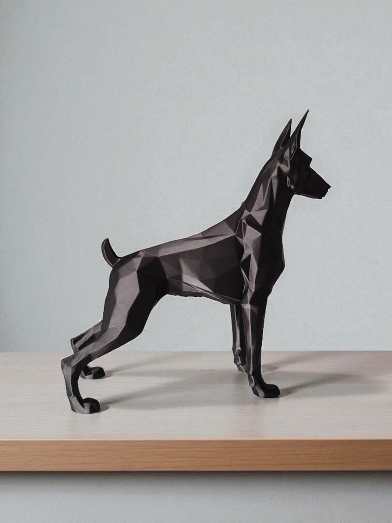 Op de afbeelding: Een zwarte geometrische sculptuur van een hond, waarschijnlijk een Dobermann, op een licht houten oppervlak. De vorm van de hond is gemaakt van hoekige vlakken, wat een moderne, abstracte uitstraling geeft. De achtergrond is effen lichtgrijs.