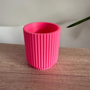 May include: A vibrant pink planter with vertical ribbed detailing. The cylindrical pot has a rounded rim and sits on a wooden surface. The bright color and textured design make it a decorative piece for indoor plants.