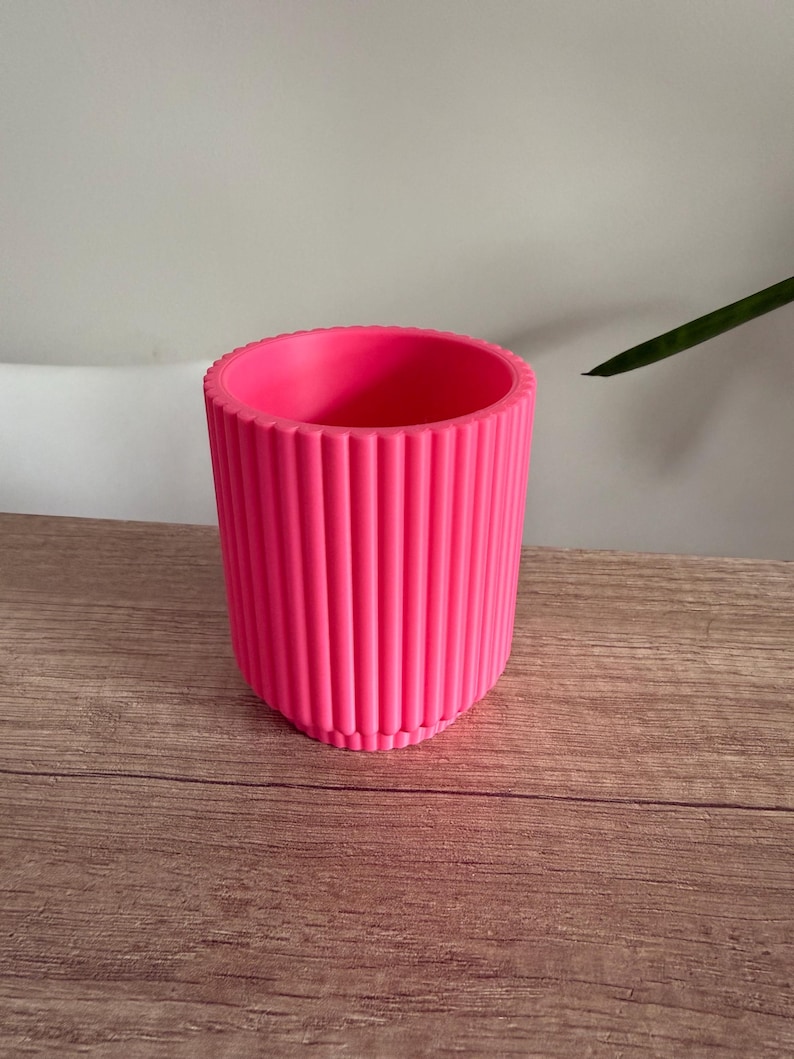 May include: A vibrant pink planter with vertical ribbed detailing. The cylindrical pot has a rounded rim and sits on a wooden surface. The bright color and textured design make it a decorative piece for indoor plants.