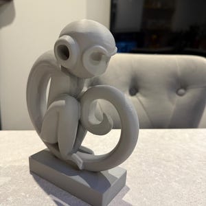 Puede incluir: Escultura de mono gris en posici&oacute;n sentada, con la cola enroscada. La escultura tiene grandes orejas circulares y est&aacute; montada sobre una base rectangular. El mono est&aacute; pintado en un color gris mate s&oacute;lido.
