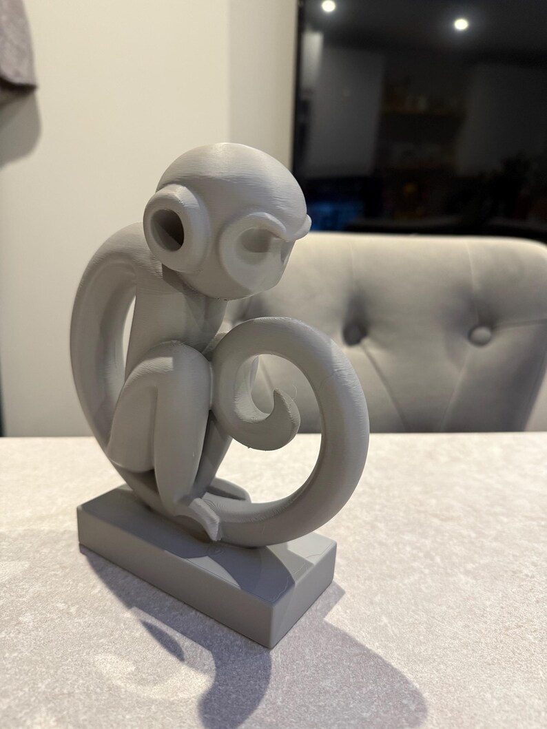 Puede incluir: Escultura de mono gris en posici&oacute;n sentada, con la cola enroscada. La escultura tiene grandes orejas circulares y est&aacute; montada sobre una base rectangular. El mono est&aacute; pintado en un color gris mate s&oacute;lido.