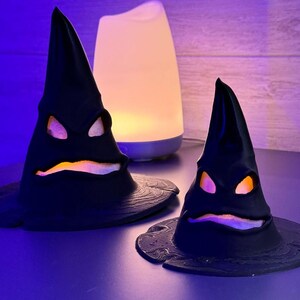 Op de afbeelding: Twee zwarte, kegelvormige decoratieve hoeden met gebeeldhouwde gezichten en gloeiende oranje en paarse lichten. De hoeden staan op een donkere ondergrond, met een witte en blauwe diffuser op de achtergrond. Halloween-thema.