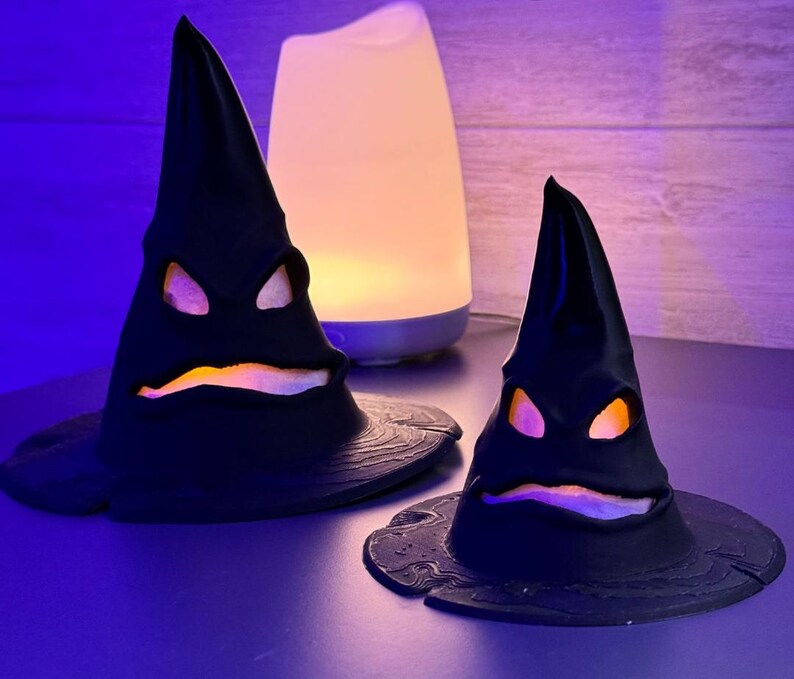 Op de afbeelding: Twee zwarte, kegelvormige decoratieve hoeden met gebeeldhouwde gezichten en gloeiende oranje en paarse lichten. De hoeden staan op een donkere ondergrond, met een witte en blauwe diffuser op de achtergrond. Halloween-thema.