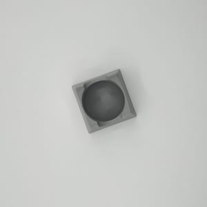 May include: A small, square, gray ashtray with a circular indentation in the center. The ashtray is made of a solid, matte material and sits on a white surface. The design is minimalist and modern.