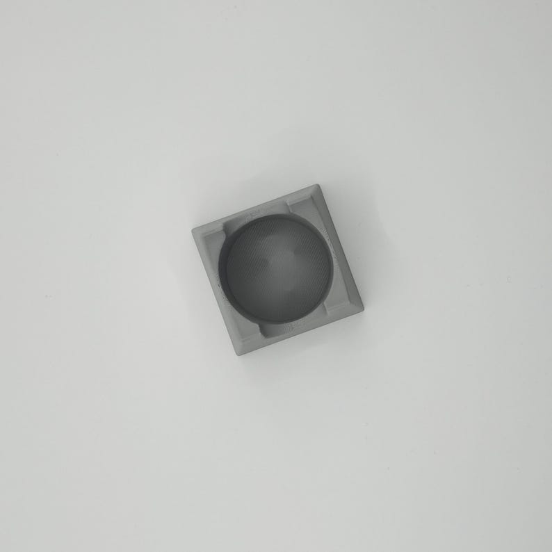 May include: A small, square, gray ashtray with a circular indentation in the center. The ashtray is made of a solid, matte material and sits on a white surface. The design is minimalist and modern.