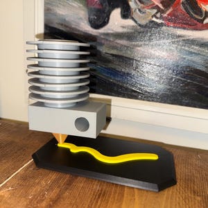 May include: A 3D printer head with a gray body and a yellow filament being extruded onto a black base. The printer head has a series of stacked, circular cooling fins. The base is a rectangular shape with a curved design.